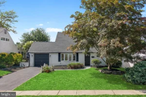 Exterior of a staged 4-bedroom Colonial home in Levittown PA with white siding, black garage door, green lawn, mature tree, and landscaped yard.