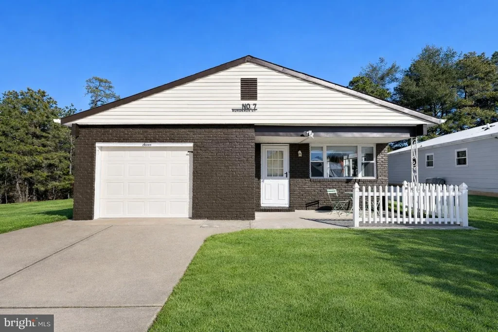 Exterior of a staged 2-bedroom ranch home in Toms River NJ with brick facade, attached garage, white picket fence, driveway, and green lawn.