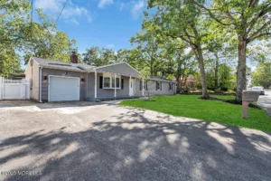 Exterior of a staged 4-bedroom Pine Lake Park ranch home in Toms River NJ with gray siding, front lawn, driveway, garage, and mature trees.